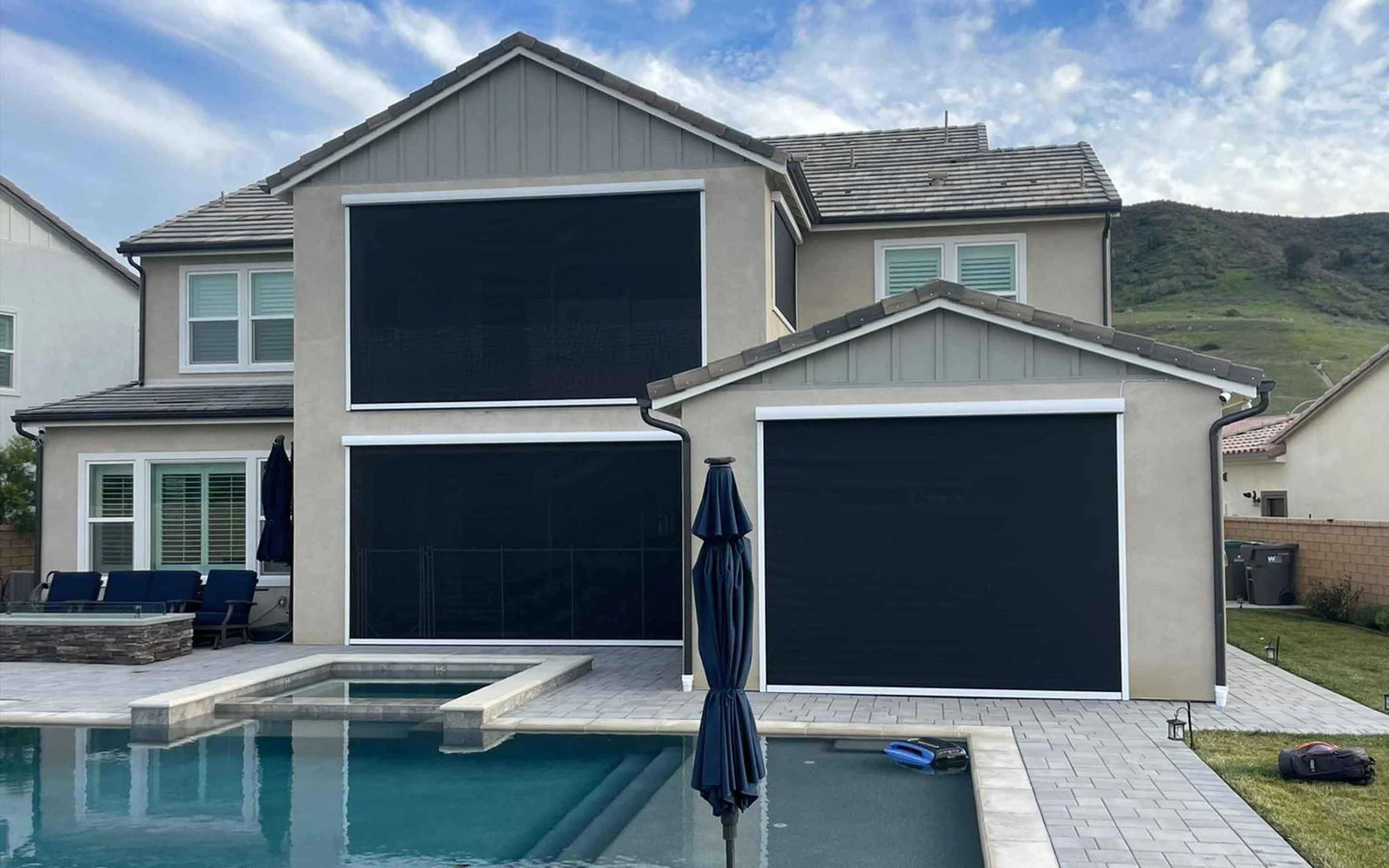 Exterior view of a luxury modern two-story home backyard featuring white-framed motorized retractable patio screens fully lowered for pool deck privacy and sun shade next to a built-in swimming pool and spa.