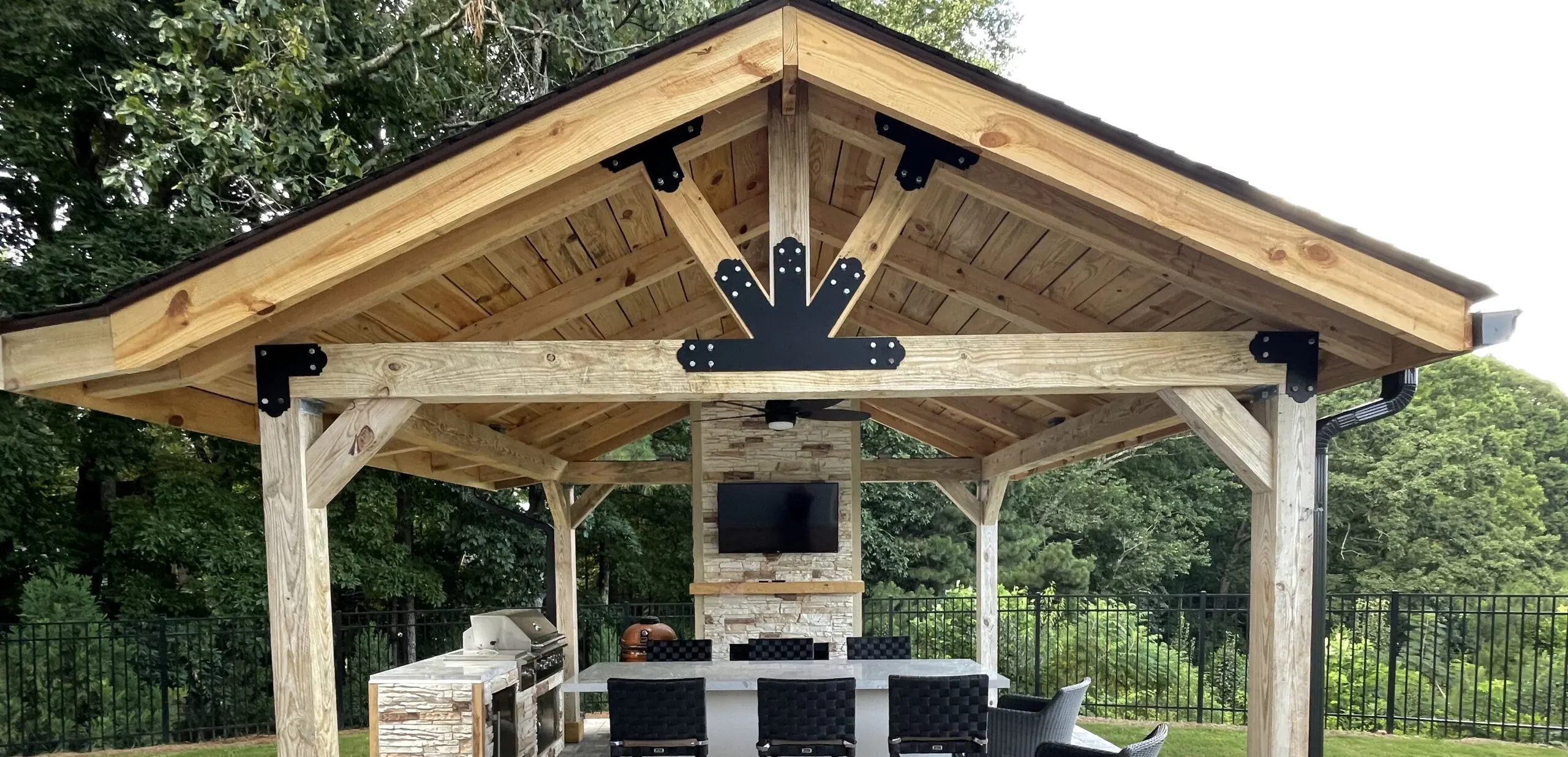 A beautiful rustic wooden pavilion pavilion with a stone fireplace, TV, and outdoor kitchen set up next to a backyard fence line.