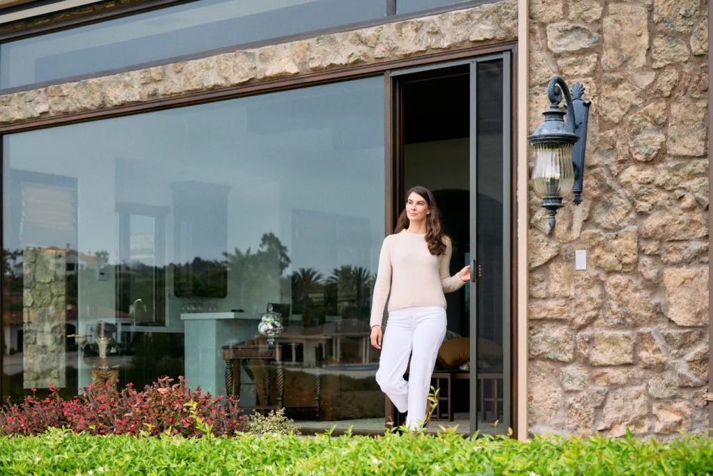 A woman standing in the large sliding glass doorway of a stone house, opening a sleek, bronze-framed retractable screen door to transition seamlessly from indoor to outdoor living space.