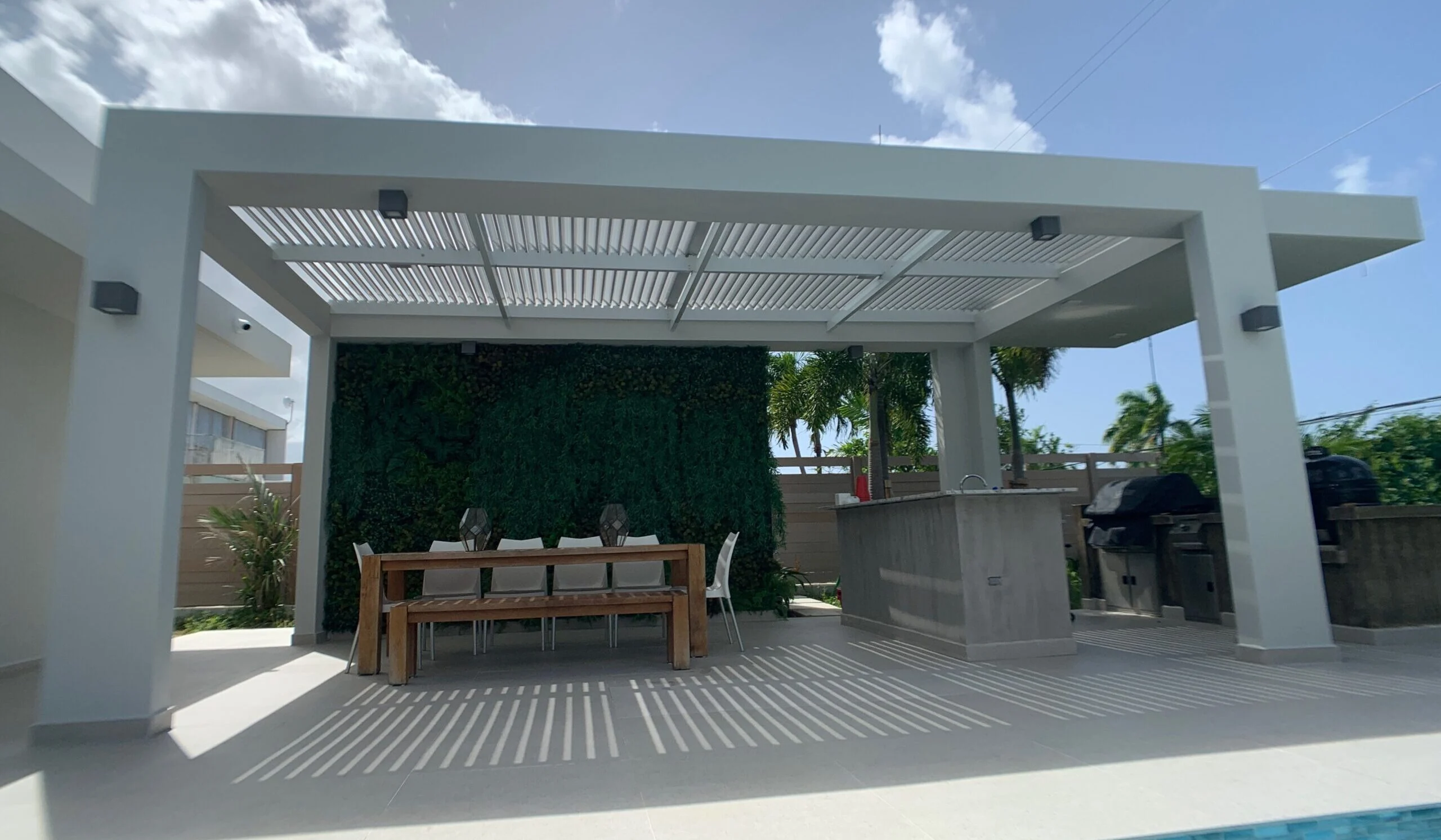 A resort-style poolside cabana featuring a large white louvered structure, a vertical plant wall backdrop, a dining area, and a barbecue cooking station under a bright blue sky.