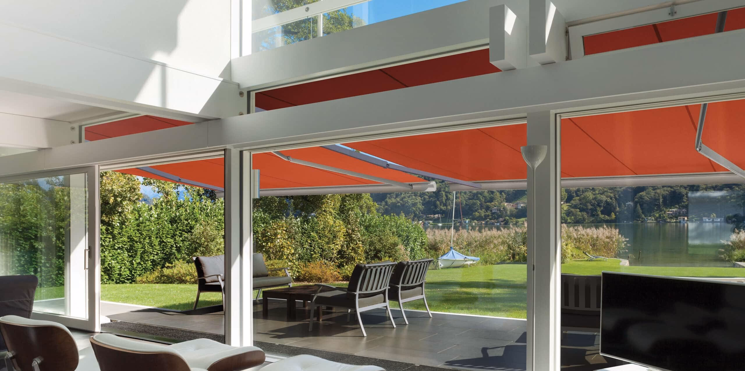 An outdoor seating area with grey chairs shaded by an orange canopy next to a green lawn, lake, and sailboat.