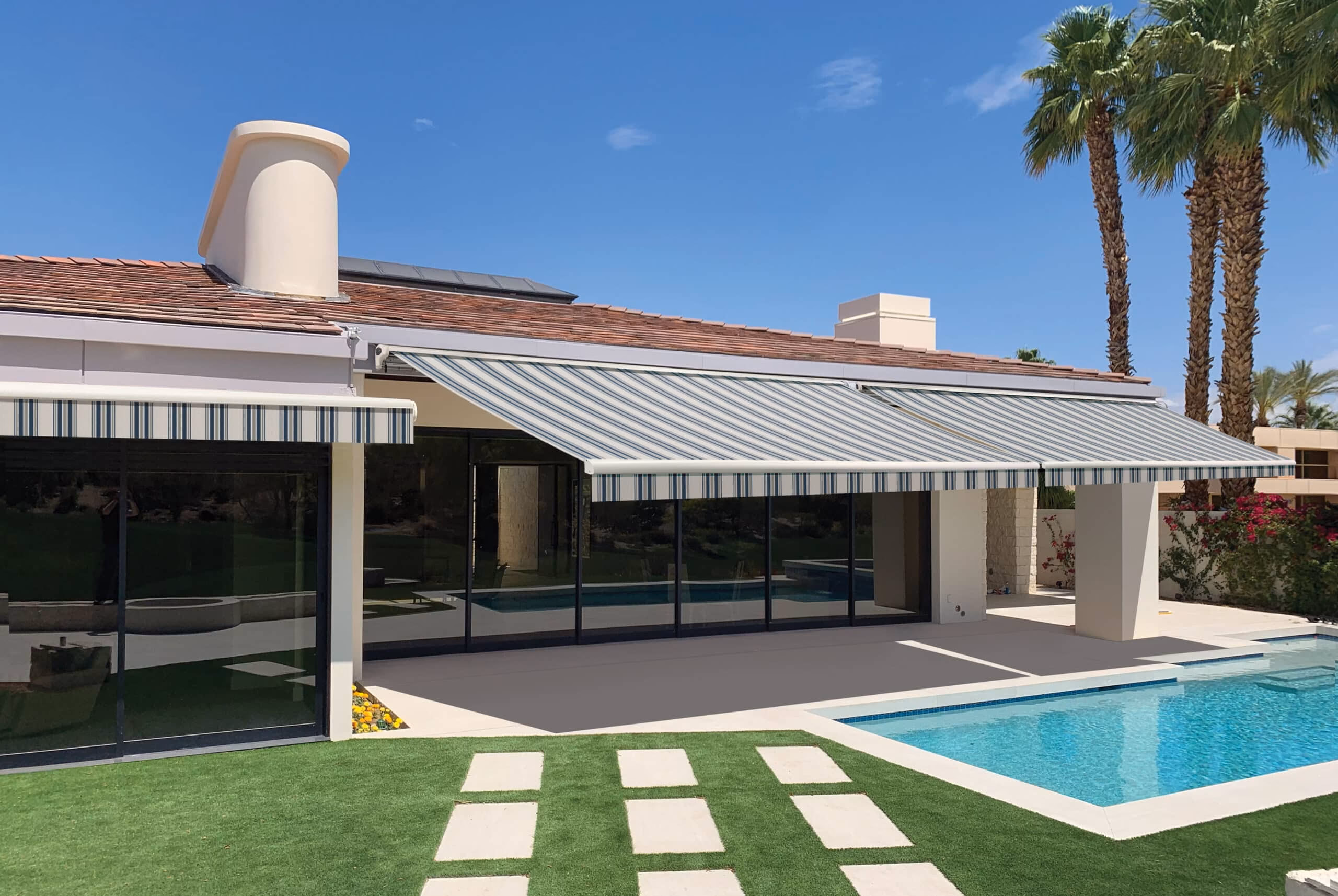 A bright backyard scene with a turquoise swimming pool, tall palm trees, artificial turf with stone steps, and extended sun awnings.