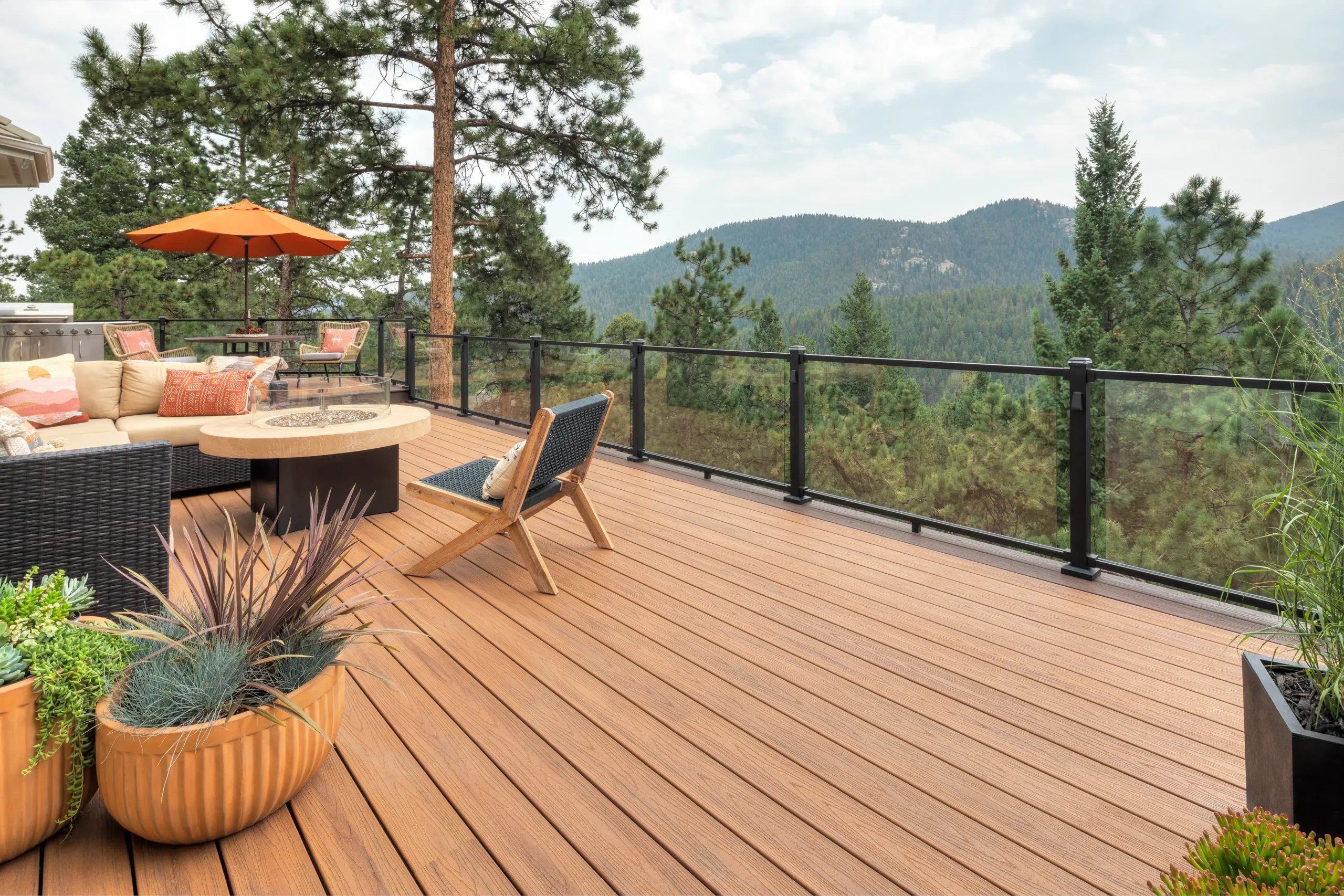 An empty area of a brown wooden deck opens up into a cozy seating arrangement with a view of green mountains in the distance.