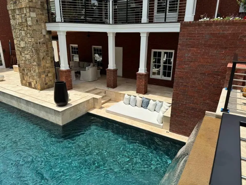A unique sunken poolside lounge layout built into a travertine stone deck, situated between a red brick house wall and a large stacked stone decorative pillar structure.