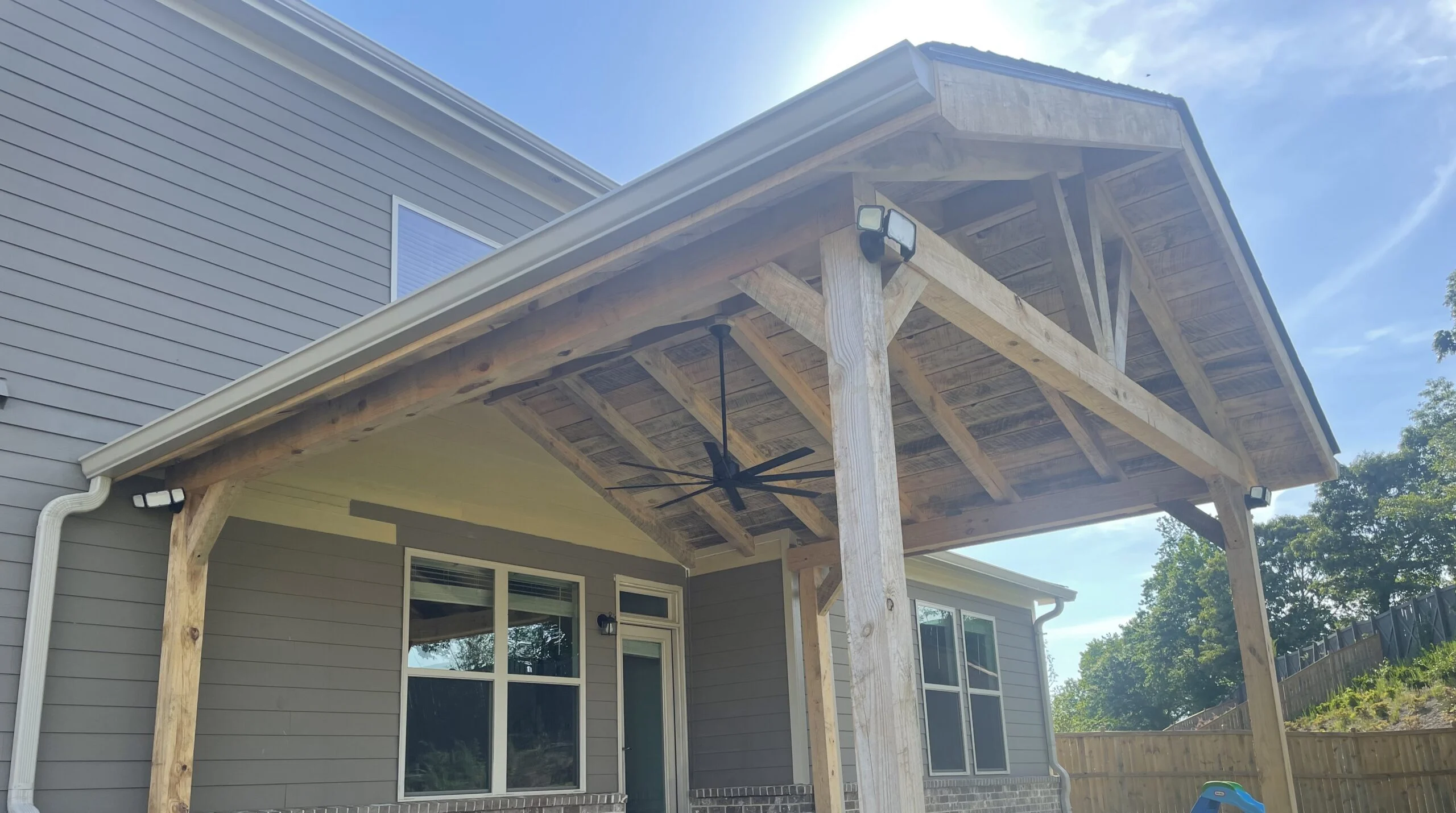 A custom-built wood post-and-beam patio cover built over a concrete porch layout, equipped with a large multi-blade black cooling fan and outdoor floodlights under a bright sky.