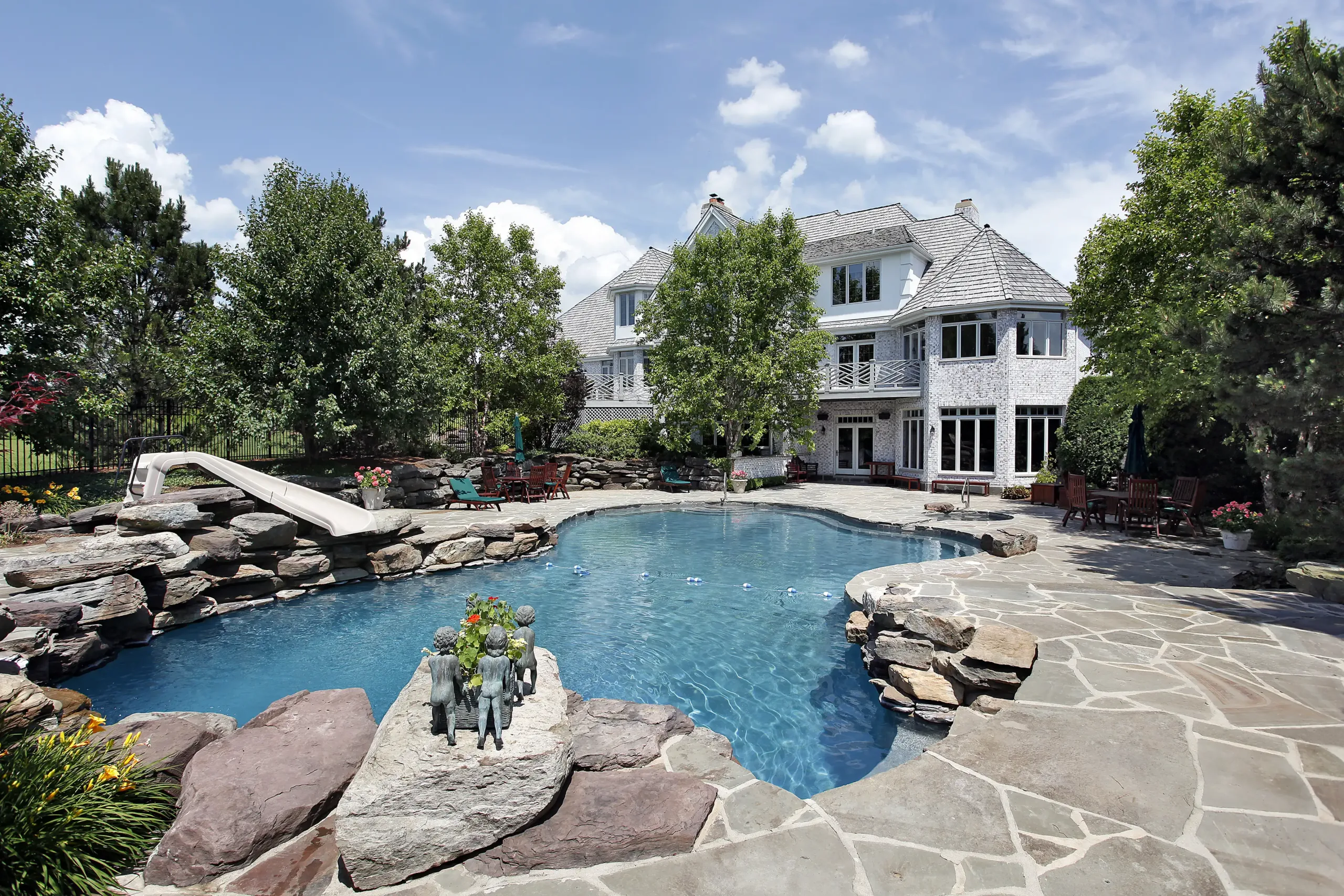 Luxury white brick home backyard featuring custom freeform pool, rock slide, and spacious flagstone stone patio.