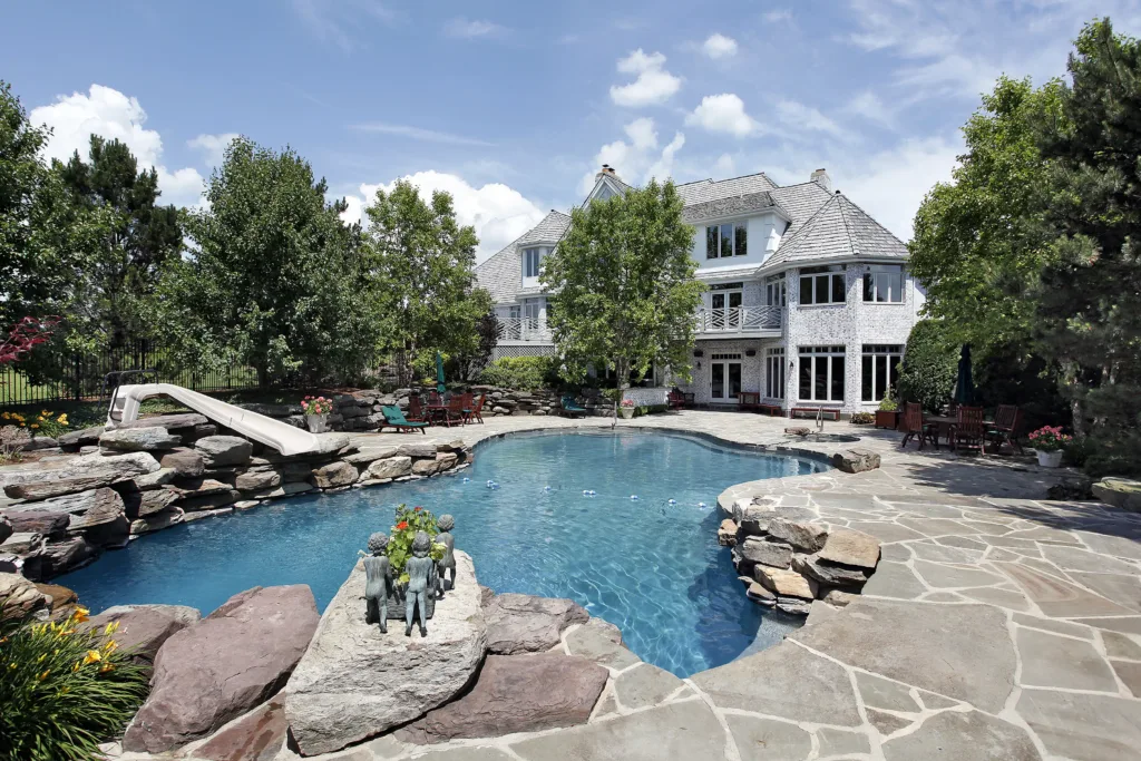Luxury backyard with freeform swimming pool, rock slide, stone patio, and large white brick house surrounded by trees.