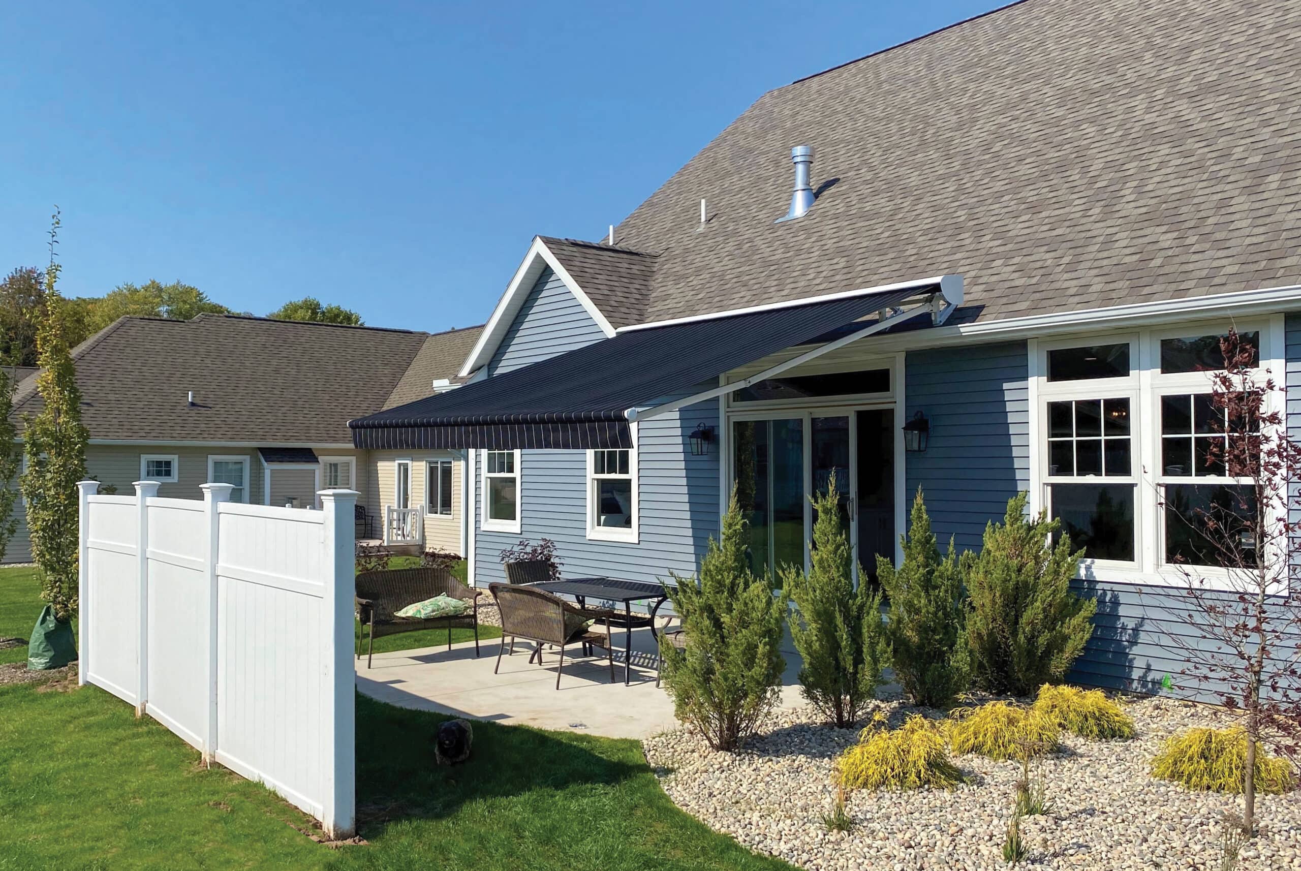 A large fabric awning mounted onto the vinyl siding of a house, providing shade over an outdoor seating area next to a rock garden.