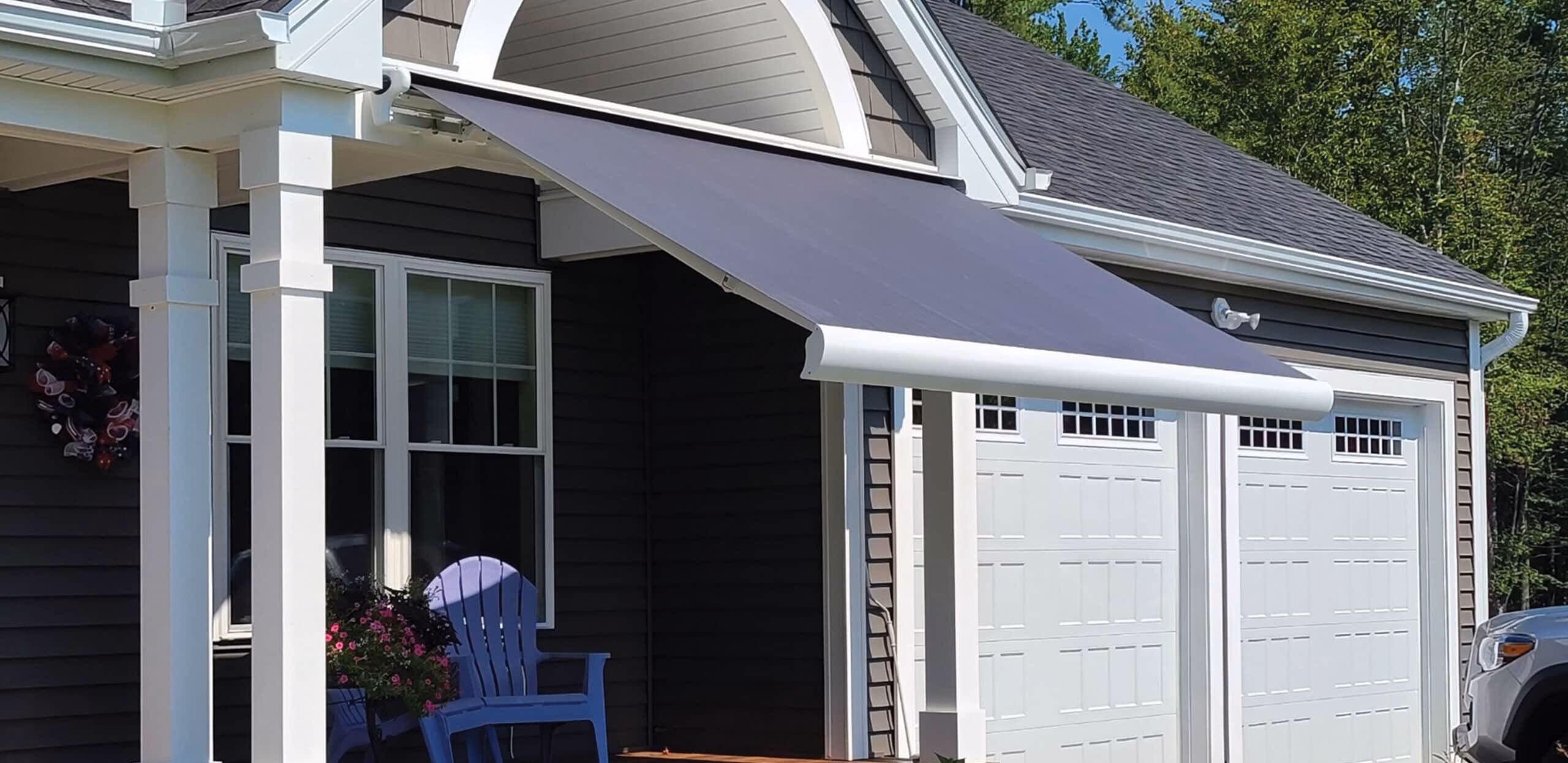 A solid gray retractable awning is extended over a dark wood porch with a blue Adirondack chair next to the garage doors.