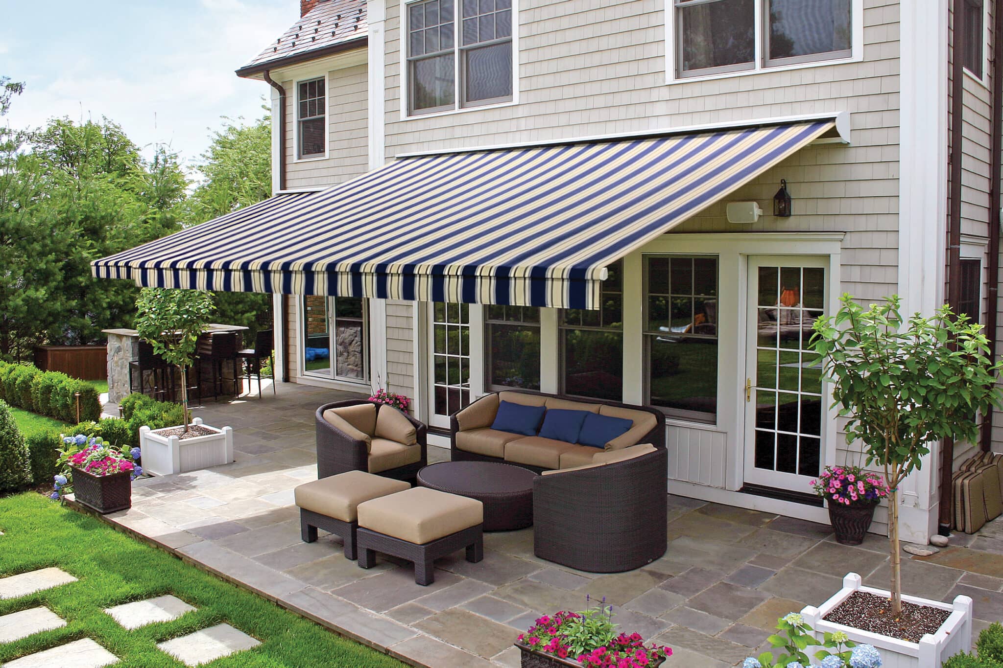 A cozy backyard patio featuring a dark brown wicker sofa set with tan cushions, blue accent pillows, potted flowers, and an attached striped sun canopy.