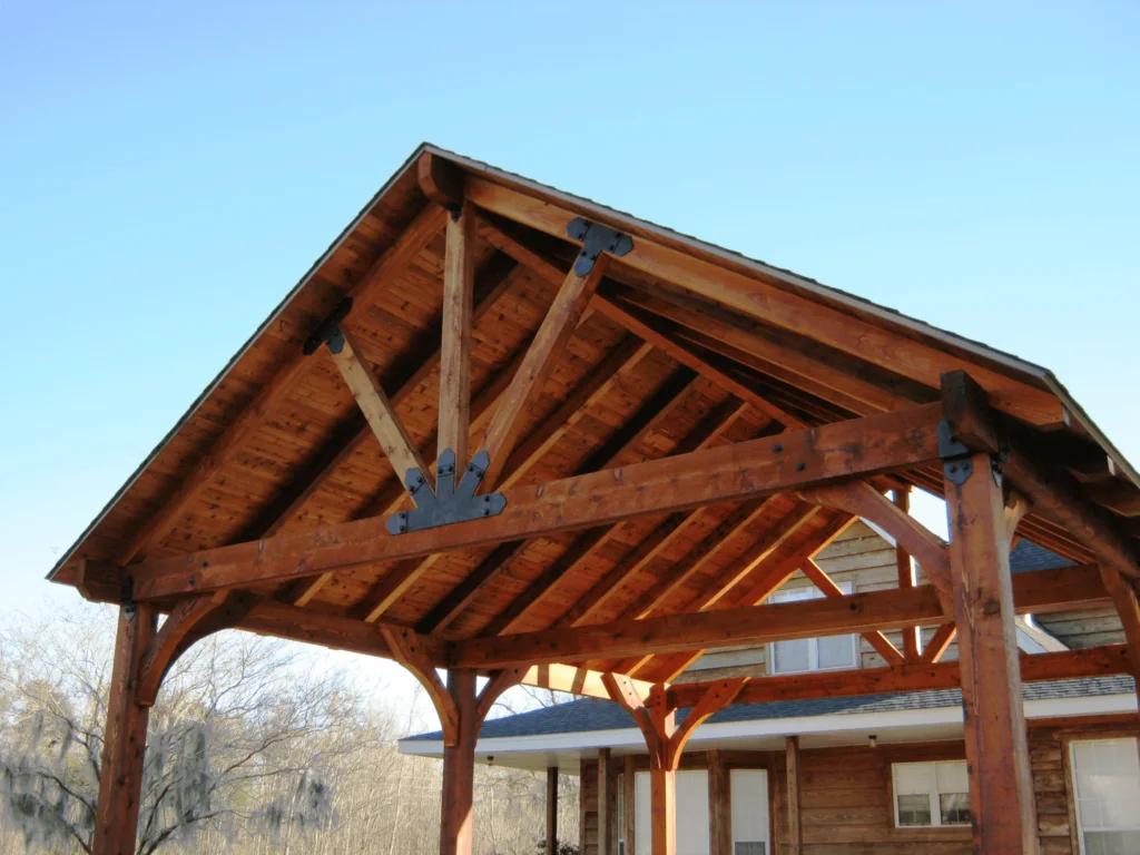 A beautifully crafted wooden pavilion gable roof structure built in front of a rustic house under a clear sky.