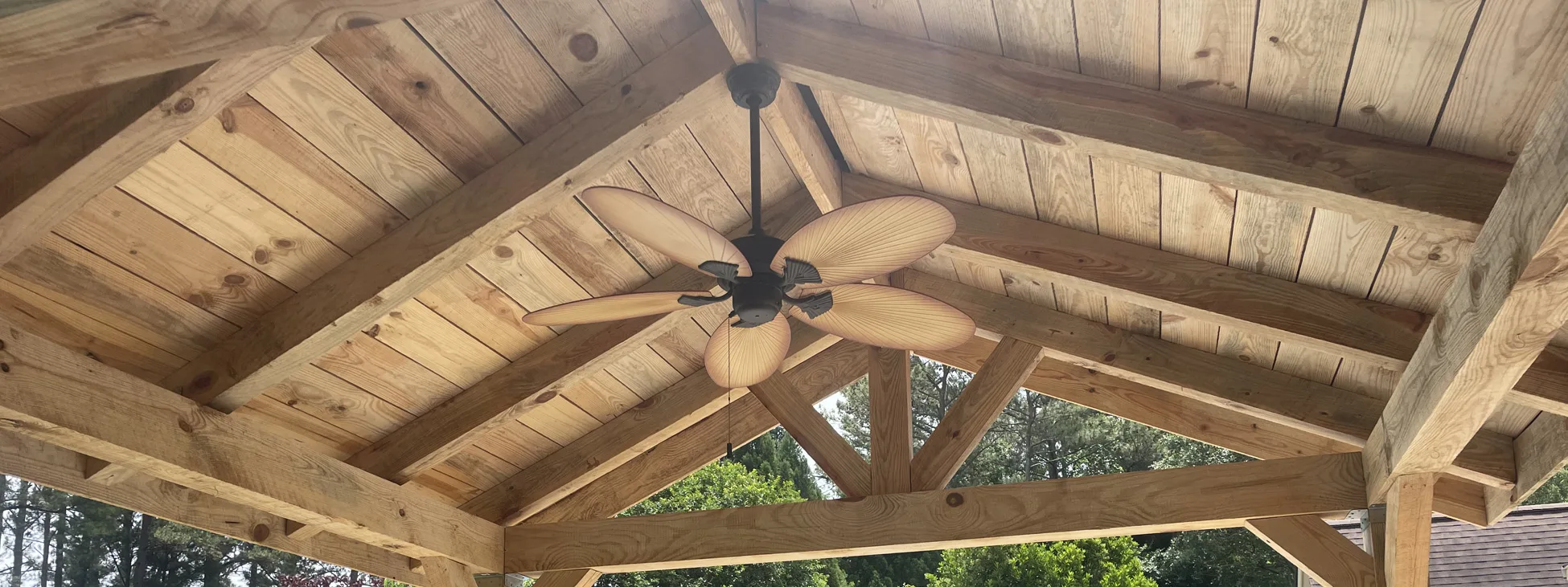 A five-blade brown ceiling fan hanging from the apex of a beautifully constructed wooden gable roof structure.
