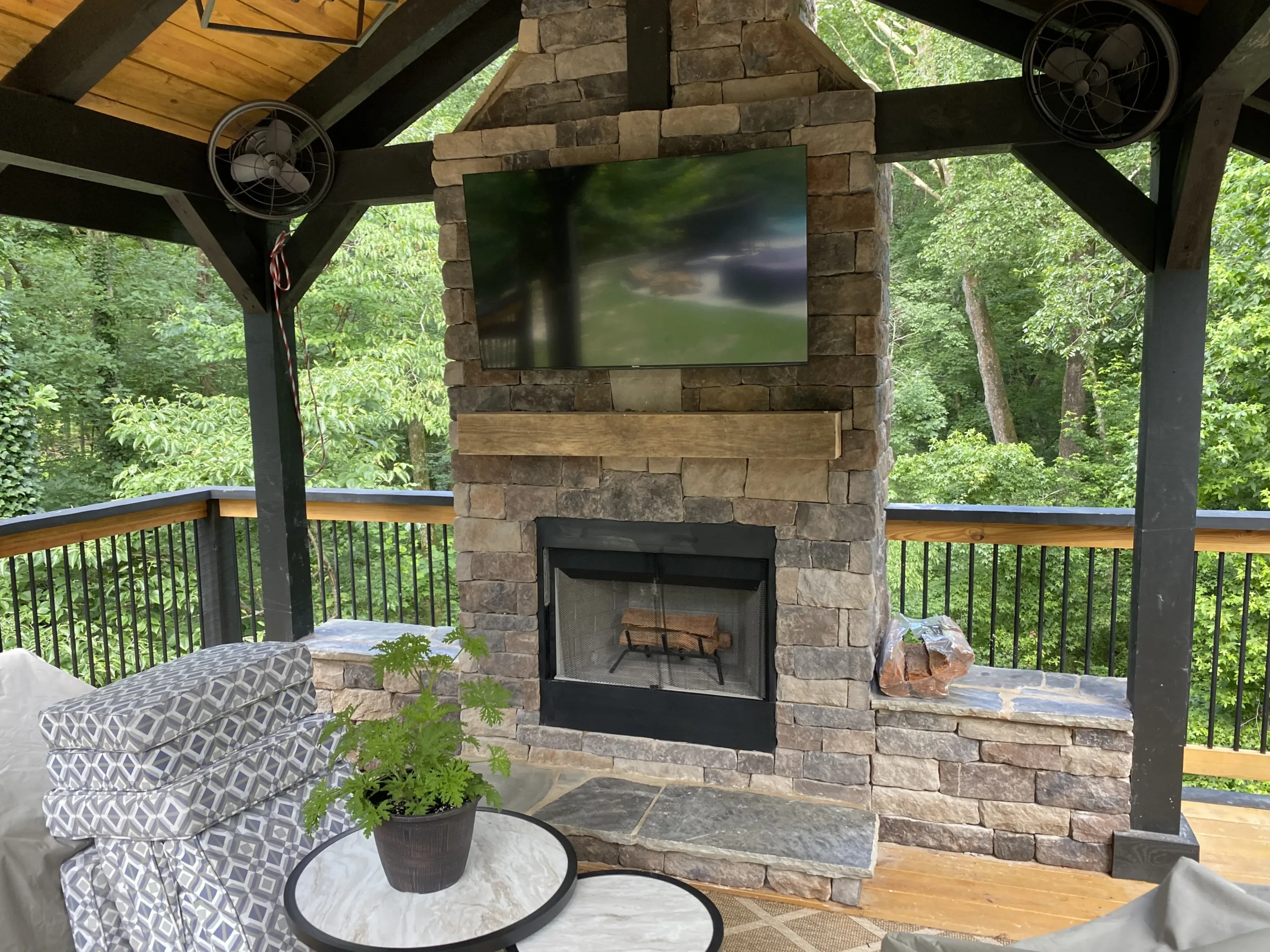 Custom backyard porch stone fireplace with integrated media wall, floating wood beam mantel, and comfortable outdoor patio lounge furniture.