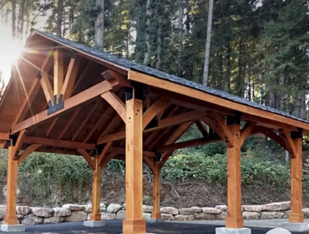 An open-air wooden timber pavilion built next to a low stone retaining wall with a dense backdrop of tall pine trees and natural forest landscaping.
